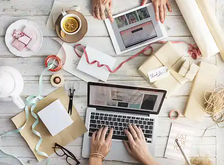 Two people work at a white wooden table with a laptop, tablet, wrapped gifts, and crafting supplies—perhaps planning a Property Owners Association event over tea.