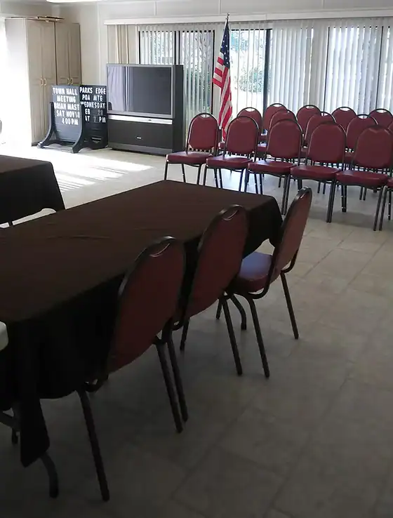 A Property Owners Association meeting room with tables covered in dark cloths, rows of red chairs, an American flag, a large TV, and a sign with community rules near large windows with vertical blinds.