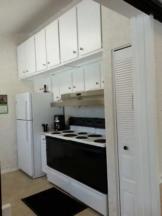 A small kitchen with white cabinets, a white refrigerator, a black and white electric stove, a range hood, coffee maker, and toaster—ideal for members of the Property Owners Association. Features include a black floor mat and folding white pantry door.