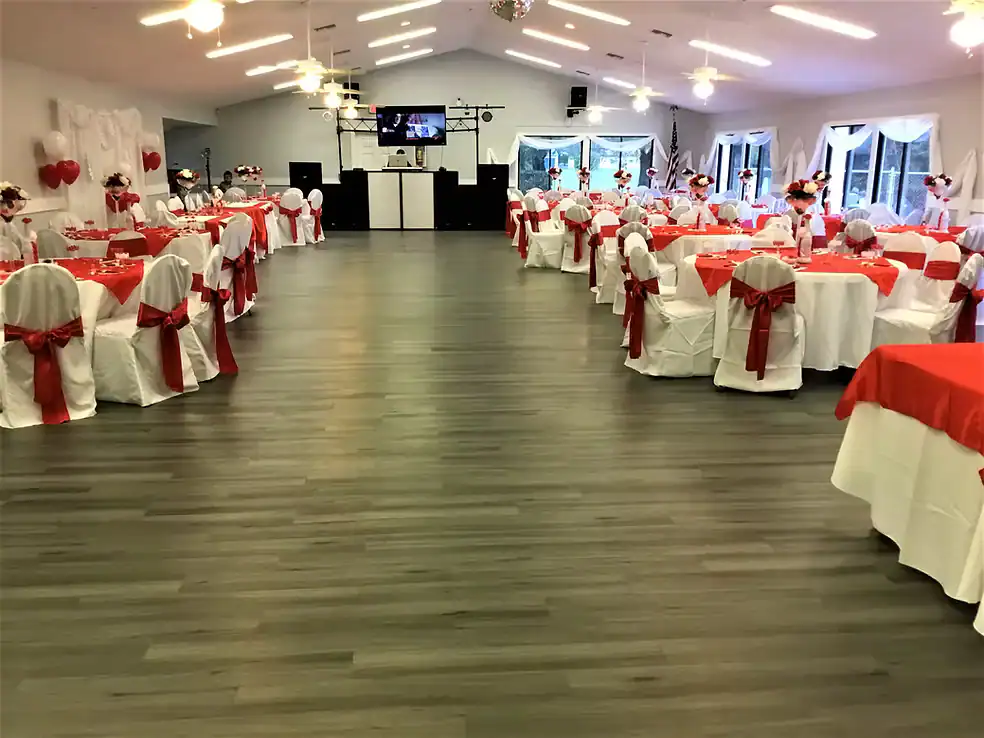 A banquet hall decorated for a Property Owners Association event with round tables covered in white tablecloths and red accents, chairs tied with red sashes, heart balloons, and a DJ setup at the front. Large windows fill the space with natural light.