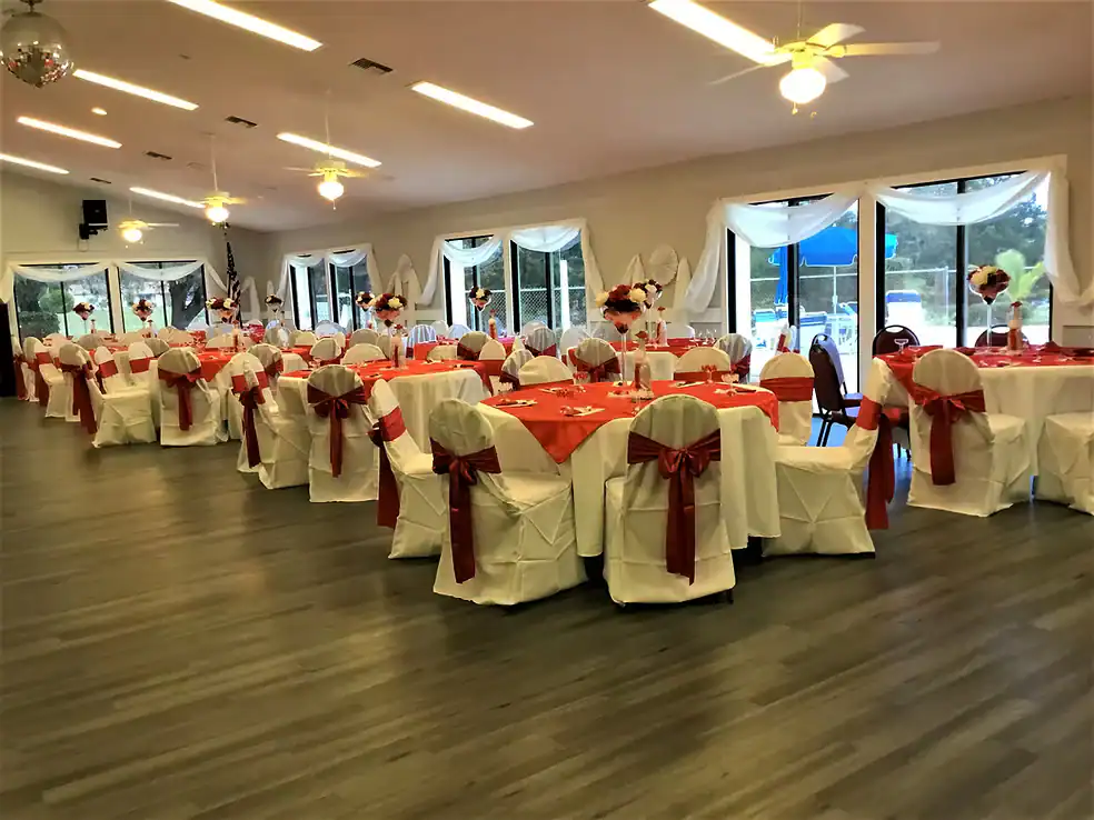 A Property Owners Association banquet hall decorated for an event with round tables covered in white cloths and red runners, chairs draped in white covers with red bows, and floral centerpieces. Large windows let in natural light.