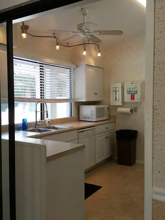 A small kitchen with white cabinets, a sink below a window with blinds, a microwave, and a wall-mounted first aid box—ideal for Property Owners Association use. Includes a paper towel dispenser, trash bin, ceiling fan, and track lights overhead.