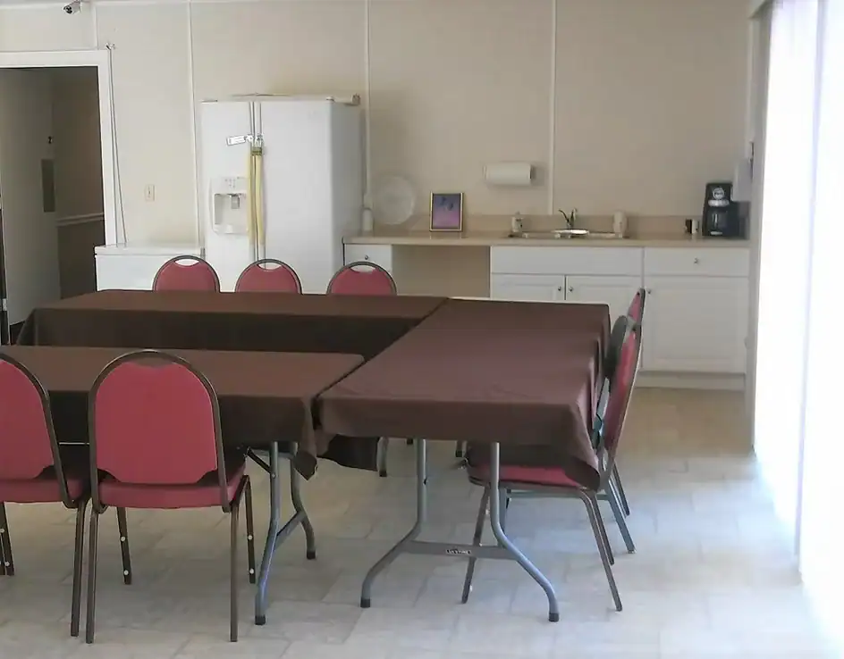 A simple room with three tables covered in brown cloths, surrounded by red chairs—ideal for Property Owners Association meetings. In the background are white cabinets, a fridge, a sink, a coffee maker, and a paper towel holder. Light streams in from the right.