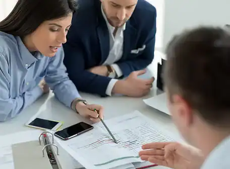 Three people in business attire sit at a desk reviewing documents and charts for the Property Owners Association. One woman points at a paper with a pen, while two men listen attentively. Phones and paperwork are scattered on the table.