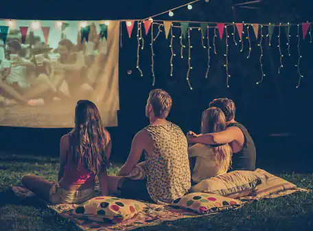Four people sit on cushions and blankets outdoors at night, enjoying a movie projected onto a screen. String lights and colorful bunting create a cozy, festive atmosphere—perfect for a Property Owners Association community event.