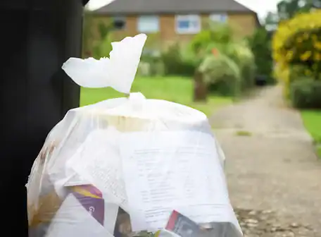 A white plastic trash bag filled with papers and discarded items sits on the ground next to a bin, near a garden path and house—an image reflecting the maintenance responsibilities of any Property Owners Association.