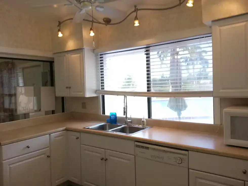 Bright kitchen with white cabinets, beige countertops, a double sink under large windows with blinds, a dishwasher, and a microwave—all within a well-maintained Property Owners Association community. Overhead lights and a ceiling fan are also visible.